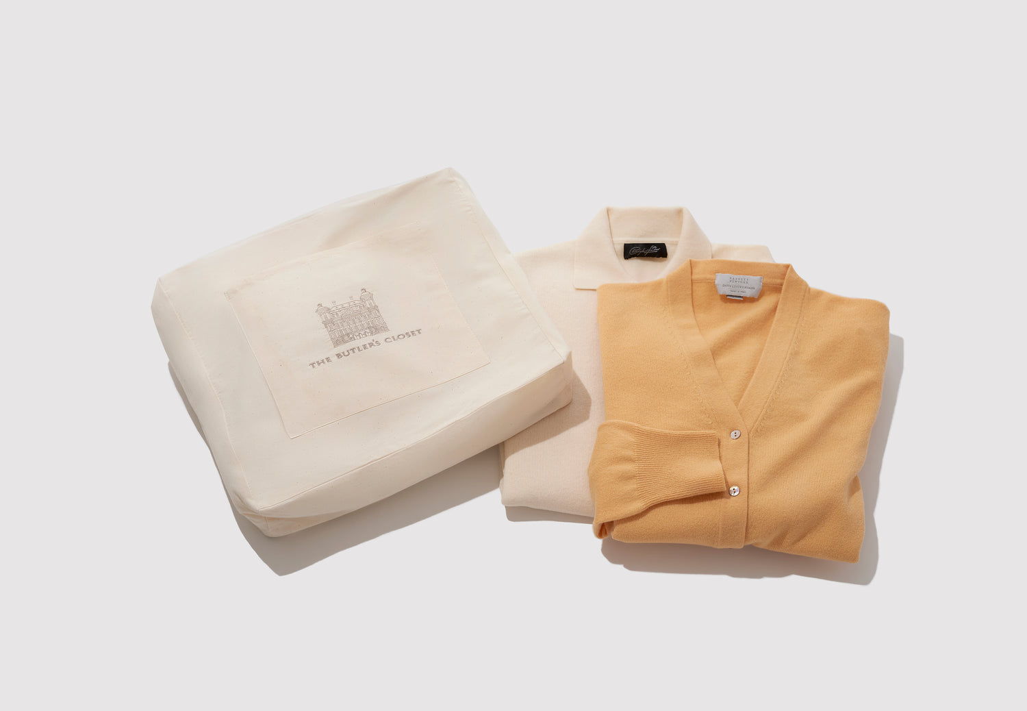 Two folded sweaters, one cream and one tan, next to a branded storage bag on a white background.