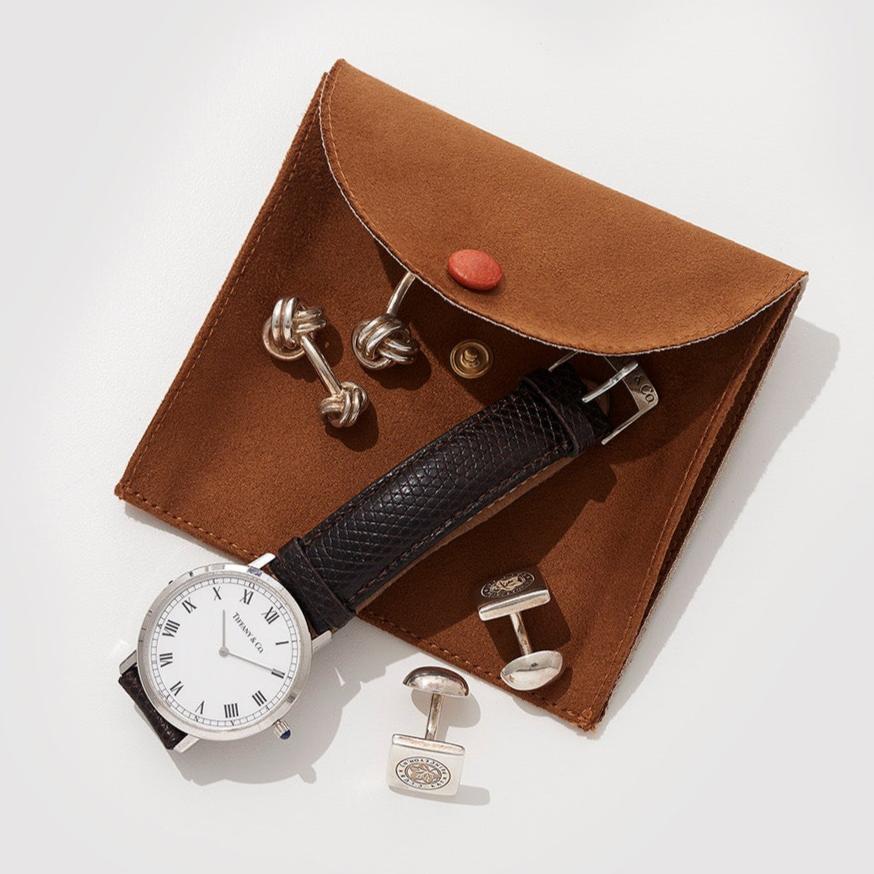 A brown leather pouch with a black watch and various silver cufflinks on a light surface.