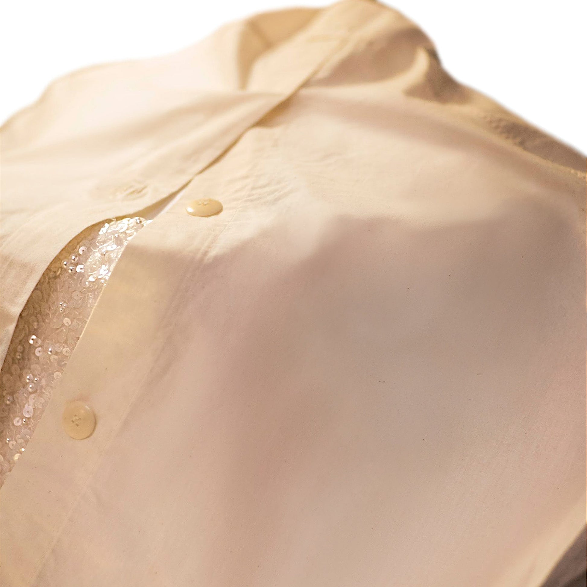 Close-up of a light beige button-up shirt with a partially visible sequin detail underneath, highlighting the fabric texture and buttons.
